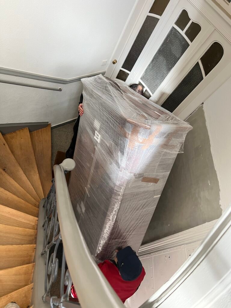 Zwei Mitarbeiter tragen ein in Folie verpacktes Möbelstück durch ein Altbau-Treppenhaus